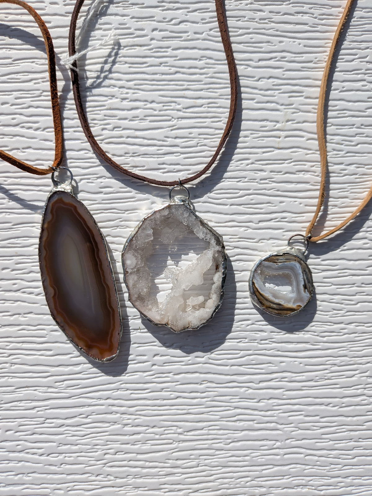Three handmade geode slice necklaces: a long brown banded pendant, an open white crystal pendant, and a small gray banded pendant.