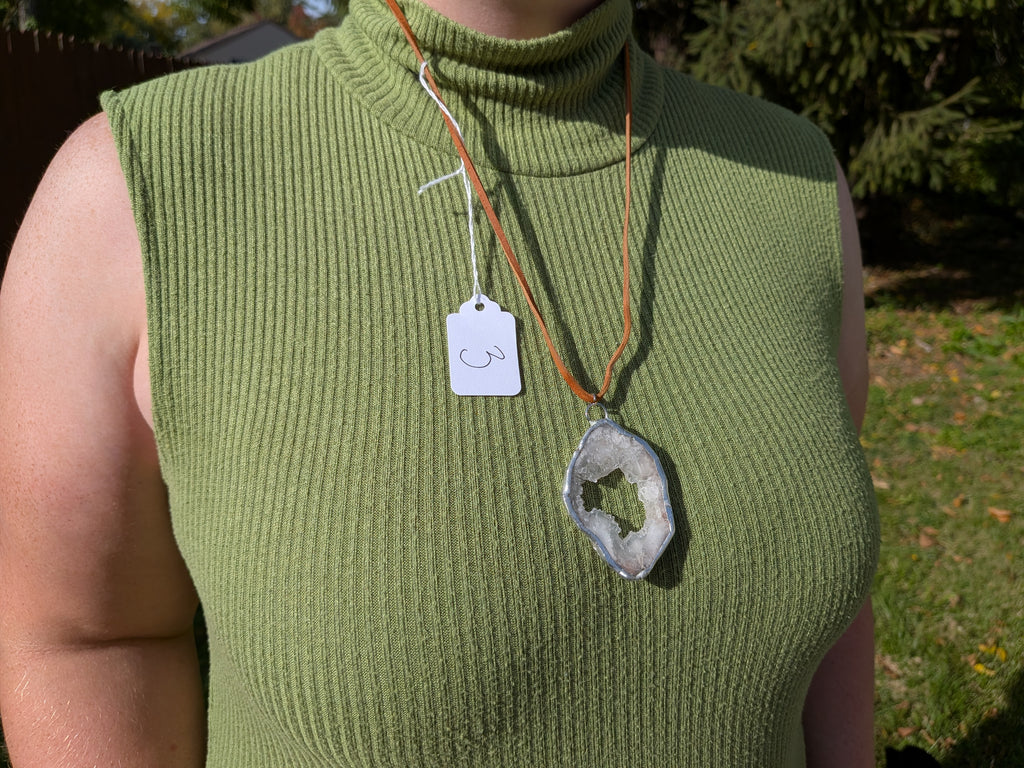 Crystal Window geode necklace No. 003 worn over a green top, showing its pale crystal-lined opening, tan cord, and pendant proportions.