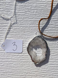 Front of Crystal Window geode necklace No. 003, with a pale crystal-lined opening, gray stone, handmade silver-tone edge, and tan cord.