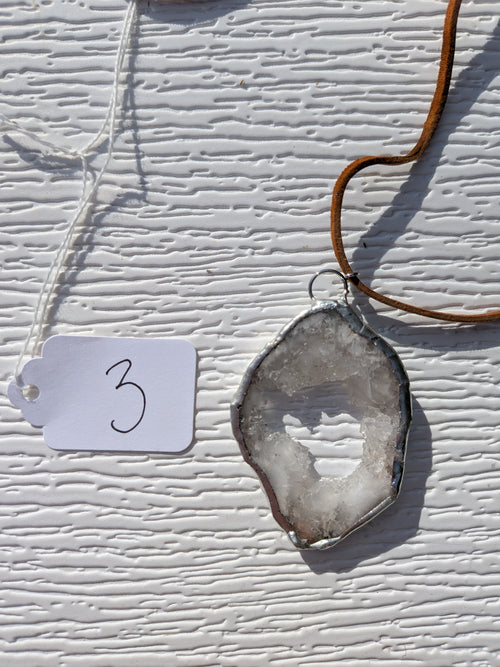 Front of Crystal Window geode necklace No. 003, with a pale crystal-lined opening, gray stone, handmade silver-tone edge, and tan cord.